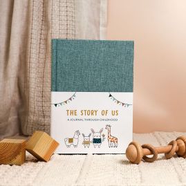 Green linen journal with white belly band, featuring illustrated bunnies, alpaca, giraffe, and bunting. Displayed on a cream rug with wooden baby toys.