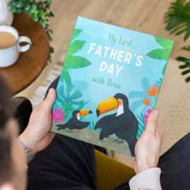 Man holding a children's book titled "My First Father's Day with Rosie," featuring two illustrated toucans and tropical plants on the cover.